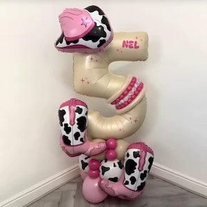 Puede incluir: Un gran arreglo de globos con el número 5 con temática de vaquera. El arreglo incluye un sombrero de vaquero rosa y blanco con estampado de vaca, botas de vaquero rosas y un número 5 beige. El nombre "NEL" está impreso en el globo.