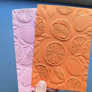 May include: Two embossed citrus-themed craft panels. One is a light pink, the other is orange. The designs feature lemons and lemon slices with leaves. The panels are held by a hand.
