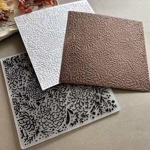 Könnte beinhalten: Drei quadratische Prägeschablonen mit einem detaillierten Blumenmuster aus Chrysanthemen und Blattzweigen. Eine Schablone ist transparent mit schwarzer Tinte, eine ist einfarbig weiß und eine ist einfarbig braun, alle mit demselben botanischen Design.