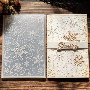 May include: A rectangular clear plastic embossing folder featuring a snowflake and dotted swirl pattern alongside a white cardstock sample embossed with the same design and a horizontal paper strip with the word Shanking in cursive script.