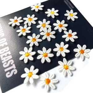 May include: White daisy flower patches with yellow centers are scattered across a dark blue card. The patches are embroidered with detailed petals. The card has the text "KING OF BEASTS" in white letters.
