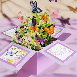 May include: A pop-up birthday card featuring a vibrant display of colorful butterflies and flowers. The card is lavender with white accents and the words "Happy Birthday" are visible on one panel. Butterflies in shades of orange, purple, pink, and blue are featured.