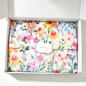 Könnte beinhalten: Ein weißer Versandkarton aus Pappe, der mit Seidenpapier ausgelegt ist, das ein buntes Aquarell-Blumenmuster in Rosa-, Gelb-, Orange- und Grüntönen aufweist. Ein rechteckiger weißer Aufkleber mit dem Text Thank you ist mittig platziert.