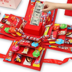 以下が含まれることがあります： キャンディーと100ドル紙幣2枚が入った赤い爆発ボックス。ボックスは「Just for You」と書かれた赤いリボンで飾られています。ボックスは、さまざまなカラフルなキャンディーやお菓子に囲まれています。