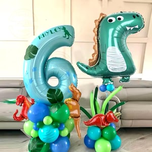 Könnte beinhalten: Ein blauer Ballon in Form der Zahl sechs mit dem Namen "Rhyley" darauf, umgeben von grünen Blättern und Dinosaurierballons. Der Ballon in Form der Zahl sechs ist Teil eines Ballonbogens mit grünen, blauen und roten Ballons.