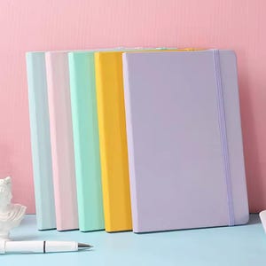 Op de afbeelding: Een verzameling van vijf notitieboeken in pastelkleuren: blauw, roze, groen, geel en lavendel. Elk notitieboek heeft een effen gekleurde kaft en een bijpassende elastische bandsluiting. De notitieboeken zijn verticaal gerangschikt.