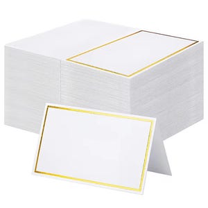 Könnte beinhalten: Ein Stapel weißer Tischkarten mit goldenem Rand. Eine Karte ist vor dem Stapel aufgestellt, ebenfalls mit goldenem Rand. Die Karten sind leer und können personalisiert werden. Ideal für Hochzeiten, Partys oder andere Veranstaltungen.