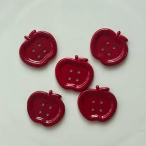 Pode incluir: Cinco botões vermelhos brilhantes em forma de maçã, cada um com quatro orifícios. Os botões estão dispostos sobre uma superfície branca. Os botões têm um detalhe de haste na parte superior. Ideais para projetos de costura e artesanato.