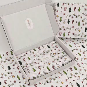 Könnte beinhalten: Ein weißer Versandkarton aus Pappe mit offenem Deckel, ausgelegt mit weißem Seidenpapier mit einem wiederkehrenden Muster aus grünen Tannenbäumen, rot-weißen Zuckerstangen, gelben Sternen und bunten Lichterketten, inklusive einem Thank You Aufkleber.