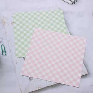 May include: Two square sheets of paper with a gingham pattern. One sheet is light green and white, and the other is pink and white. The papers are on a white, distressed wooden frame.