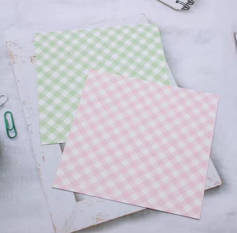 May include: Two square sheets of paper with a gingham pattern. One sheet is light green and white, and the other is pink and white. The papers are on a white, distressed wooden frame.