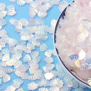 Op de afbeelding: Iriserende schelpvormige knutselversieringen verspreid op een lichtblauwe ondergrond, met enkele in een wit en blauw omrande kom. De schelpen hebben een parelmoerglans, die verschillende kleuren reflecteert. Ideaal voor knutsel- en decoratieprojecten.