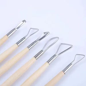 Könnte beinhalten: Ein Set aus sechs Töpferwerkzeugen mit Holzgriffen und Edelstahlköpfen. Die Werkzeuge haben unterschiedlich geformte Köpfe, darunter Schlaufen und abgewinkelte Klingen, zum Formen und Schnitzen von Ton. Die Werkzeuge sind auf einer weißen Oberfläche angeordnet.