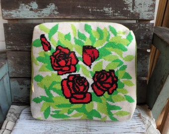 May include: A white pillow with a green and white floral design. The pillow features red roses with black outlines and green leaves. The pillow is sitting on a white wooden surface.