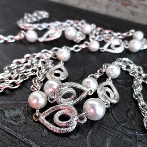 May include: A silver chain necklace with white pearls and a swirl design. The necklace is on a black textured surface.