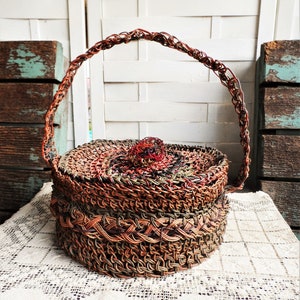 May include: A woven wicker basket with a lid and a handle. The basket is brown, green, and red. It is sitting on a white lace doily.