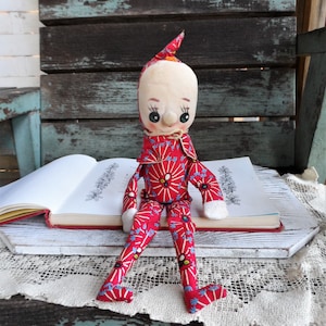 May include: A red and white patterned fabric doll with a pointed hat and large eyes. The doll is sitting on a book with a white lace doily underneath.