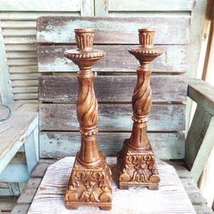 May include: A pair of gold-toned metal candle holders with ornate designs. The candle holders have a twisted, fluted design and a square base with intricate details.