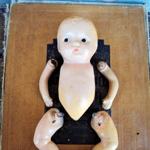 May include: A vintage porcelain doll in pieces, including the head, torso, and two legs. The doll is painted in a light skin tone with blue eyes and a pink mouth. The doll is displayed on a brown leather background.