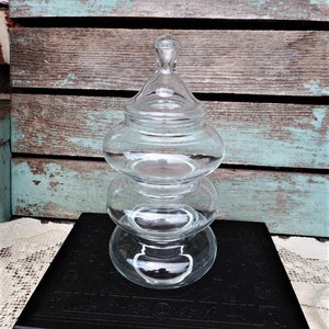 May include: A clear glass apothecary jar with a lid, featuring a stacked, rounded design. The jar sits atop a black book with embossed detailing, against a backdrop of weathered wooden planks with turquoise paint.