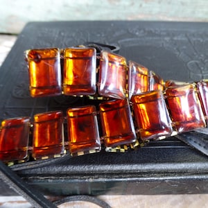 May include: A vintage bracelet featuring rectangular amber-colored glass beads. The beads are connected by small gold-toned metal links and a black cord. The bracelet is displayed on a textured black surface.