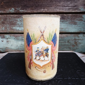 May include: A cylindrical glass with a textured, frosted appearance. The glass features a central emblem with figures in historical attire, flanked by crossed rifles and American flags. The design is in shades of beige, red, blue, and green.