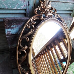 May include: A gold-toned oval mirror with an ornate, scrolled frame. The mirror reflects a wooden railing and a portion of a car.