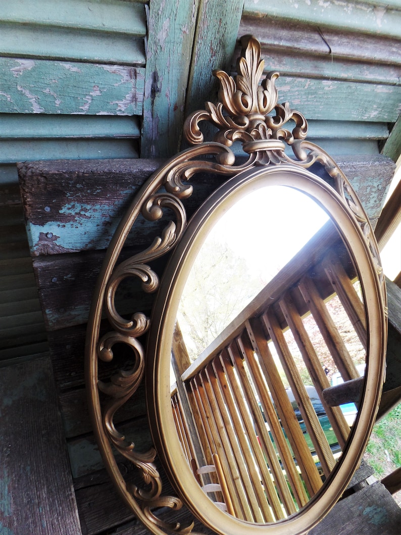May include: A gold-toned oval mirror with an ornate, scrolled frame. The mirror reflects a wooden railing and a portion of a car.