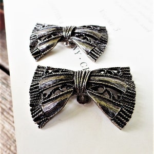 May include: Two silver-toned bow-shaped brooches with intricate filigree designs. The brooches have a textured, almost woven, appearance.