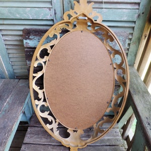 May include: A gold oval mirror with a decorative ornate frame. The mirror is hanging on a wooden wall.