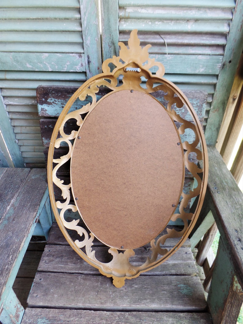 May include: A gold oval mirror with a decorative ornate frame. The mirror is hanging on a wooden wall.