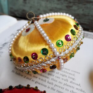 May include: A gold Christmas ornament with a satin finish and a beaded trim. The ornament is decorated with colorful sequins and a silver ribbon.