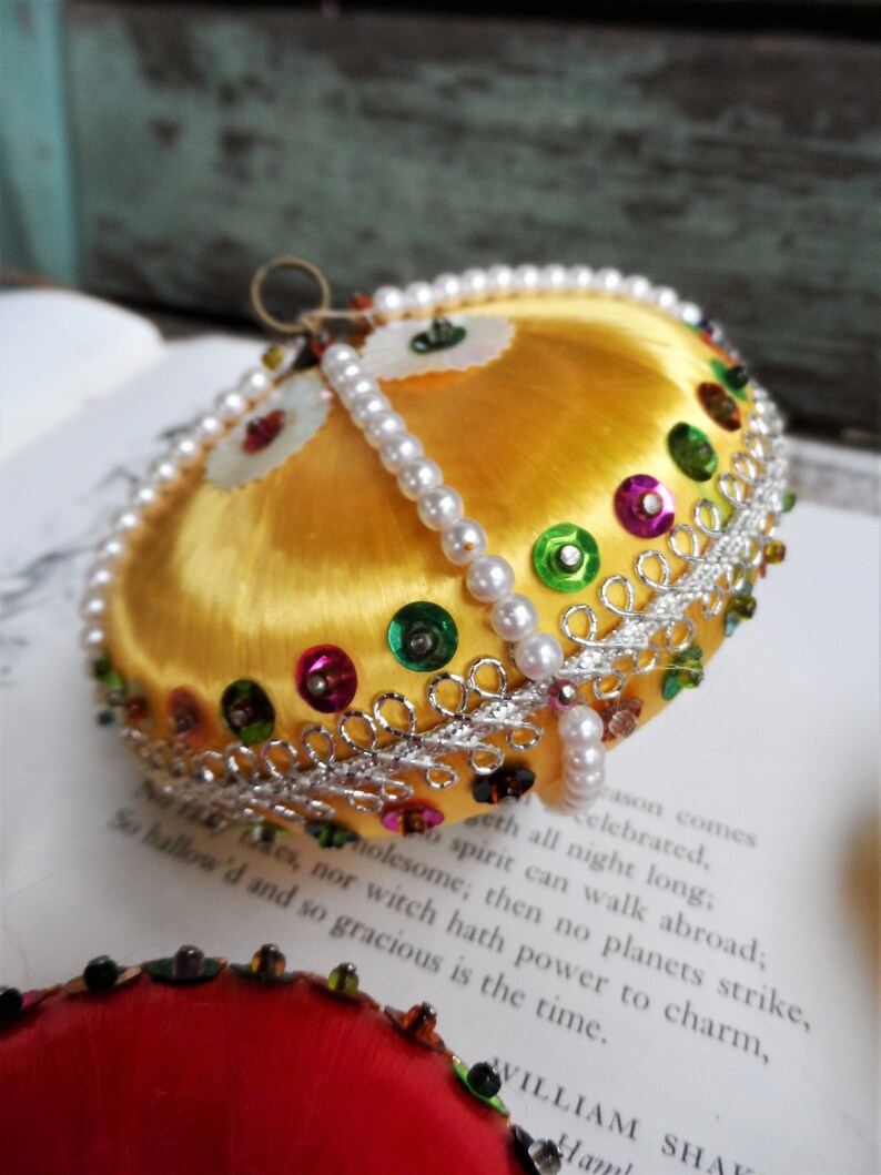 May include: A gold Christmas ornament with a satin finish and a beaded trim. The ornament is decorated with colorful sequins and a silver ribbon.
