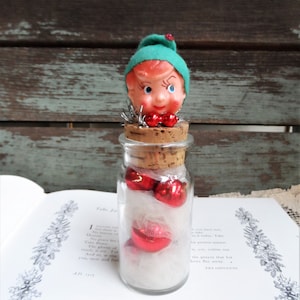 May include: A small glass jar with a cork lid filled with red ornaments and a green elf figurine on top. The jar is sitting on a book with text.