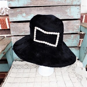May include: A black wide-brimmed hat with a large rhinestone embellishment in the shape of a rectangle.