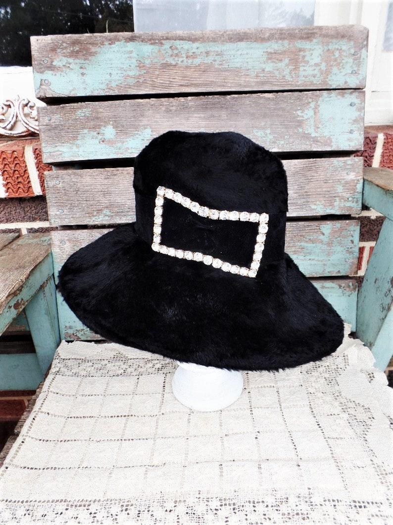 May include: A black wide-brimmed hat with a large rhinestone embellishment in the shape of a rectangle.