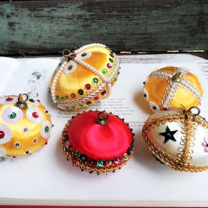 May include: Five vintage Christmas ornaments made of satin fabric and decorated with sequins, beads, and gold trim. The ornaments are in various colors, including yellow, red, and white.