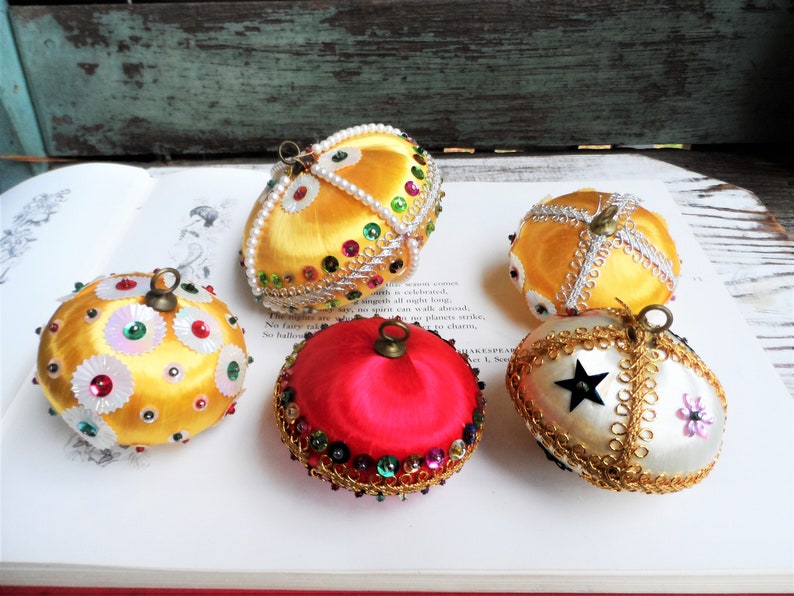 May include: Five vintage Christmas ornaments made of satin fabric and decorated with sequins, beads, and gold trim. The ornaments are in various colors, including yellow, red, and white.