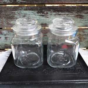 May include: Two clear glass jars with lids, sitting on a black book. The jars have a square shape and a textured design.