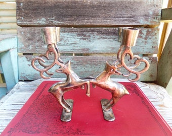 May include: A silver candle holder featuring two stylized deer with antlers, facing each other. The deer are holding up two candle cups.