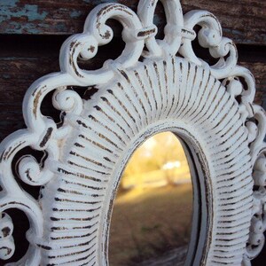 May include: White ornate oval mirror with a distressed finish. The mirror has a decorative frame with a raised design. The mirror is reflecting a blurry image of trees and a sky.
