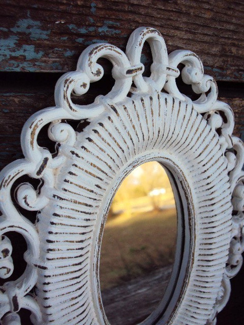 May include: White ornate oval mirror with a distressed finish. The mirror has a decorative frame with a raised design. The mirror is reflecting a blurry image of trees and a sky.