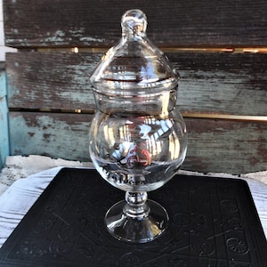 May include: A clear glass jar with a domed lid and a wide base. The jar is sitting on a black book with an ornate design.