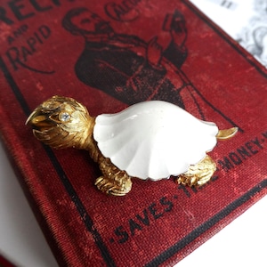 May include: A gold-toned turtle brooch with a white shell and a small clear gemstone on its head. The brooch is set on a red book with the words "READY RECKONER" visible.
