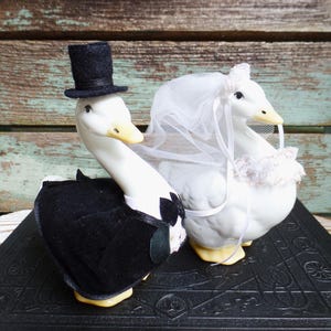 May include: Two white ceramic goose figurines dressed for a wedding. One goose wears a black velvet tuxedo jacket with a bow tie and a black felt top hat. The other goose wears a white tulle veil with a floral headpiece and a white lace collar.