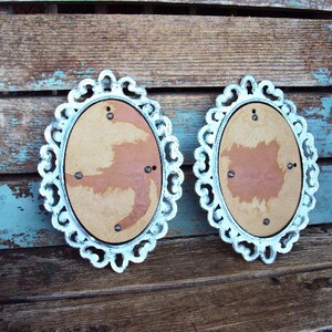 May include: Two white oval picture frames with ornate edges. The frames are made of wood and have a distressed white paint finish. The frames are hanging on a wooden wall.