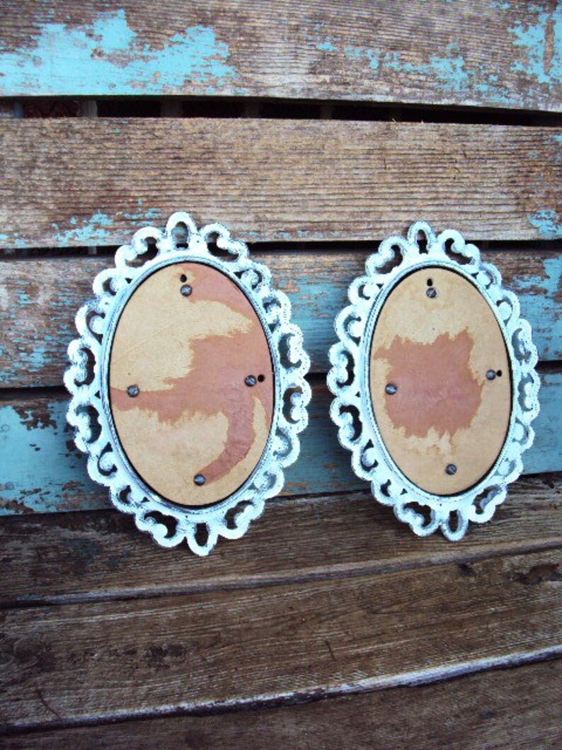 May include: Two white oval picture frames with ornate edges. The frames are made of wood and have a distressed white paint finish. The frames are hanging on a wooden wall.
