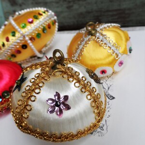 May include: Three vintage Christmas ornaments with gold and white satin fabric, gold trim, and sequins. The ornaments are round and have a traditional design.