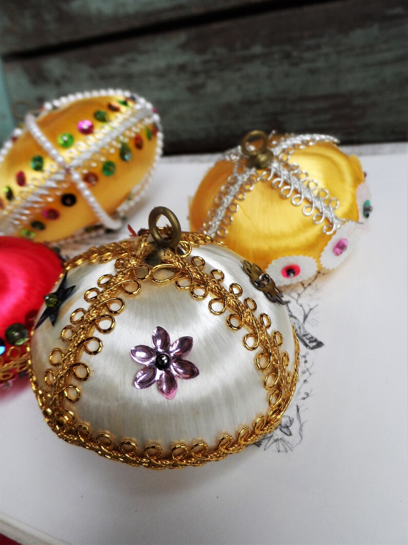 May include: Three vintage Christmas ornaments with gold and white satin fabric, gold trim, and sequins. The ornaments are round and have a traditional design.