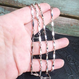 May include: A silver-toned necklace with an elongated oval link design. The necklace is composed of multiple strands, each featuring a series of symmetrical links connected by small circular rings. The links have a polished, reflective surface, and the necklace is displayed against a neutral background.